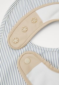 Bavoir rayé avec bord beige, présentant un design à col rond. Deux boutons pression et une texture lisse sur le tissu.