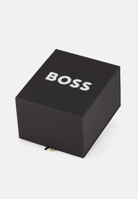 Schwarze rechteckige Box mit matter Oberfläche. Auf der Oberseite befindet sich fett gedruckter weißer Text mit der Aufschrift „BOSS“ und ein kleines, mehrfarbiges Banddetail am Boden.