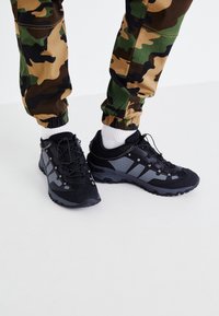 Zwarte en grijze wandelschoenen met gemusterde veters, gedragen met witte sokken en camouflagebroek op een witte vloer.