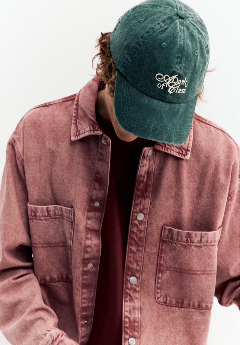 Person trägt eine dunkelgrüne Kappe mit der Aufschrift "Bash of Class" und eine verblasste rote Hemdjacke, den Kopf gesenkt, vor schlichtem weißem Hintergrund.