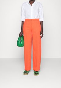 Pantaloni in lino arancioni con vita alta e gamba diritta, abbinati a una camicia bianca con bottoni e una borsa verde strutturata. Sandali verdi in evidenza.