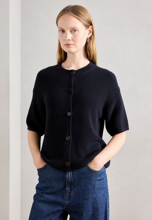 Woman wearing a navy short-sleeve buttoned cardigan and blue jeans, standing with hands in pockets against a plain light background.