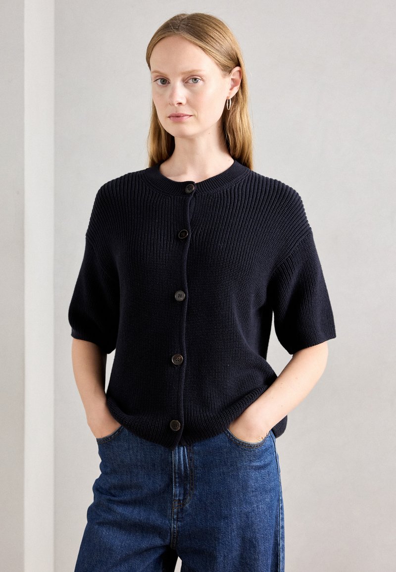 Femme portant un cardigan boutonné à manches courtes bleu marine et un jean bleu, se tenant avec les mains dans les poches devant un fond clair uni.