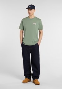 Zelené crewneck tričko s logem "Dickies", doplněné tmavě modrými volnými džínami a béžovými pracovními botami. Doplňkem je tmavě modrá kšiltovka.