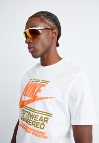 Sportske naočale s tirkiznim i narančastim lećama, bijelim okvirom i reflektirajućim detaljima, nošene uz bijelu majicu s šarenim Nike brendiranjem.