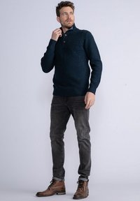 Pulover tricotat navy cu guler înalt și închidere cu trei nasturi, asociat cu blugi gri închis și cizme maro cu șiret.