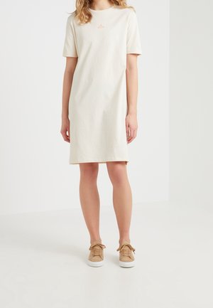 Kvinde iført en knælængde, kortærmet beige kjole med et lille bøjleikon på brystet og beige snørestøvler.