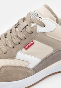 Sneaker beige e grigio con pannelli in suede e mesh, dettagli bianchi, lacci beige e etichetta rossa del logo Levi's sul lato. Suola in gomma visibile.