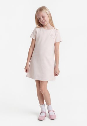 Jeune fille aux cheveux blonds souriante, portant une robe rose clair, des chaussettes blanches et des chaussures roses, debout sur un fond blanc.
