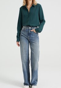 Mörkgrön långärmad knäppskjorta ihop med hög midja ljusblå raka jeans och vita sneakers. Enkelt, funktionellt design.