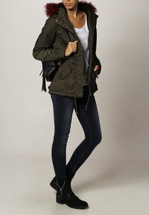 Una mujer lleva una parka verde con capucha de piel roja, una camisa blanca, jeans ajustados oscuros, botas de tobillo negras y lleva una mochila negra.