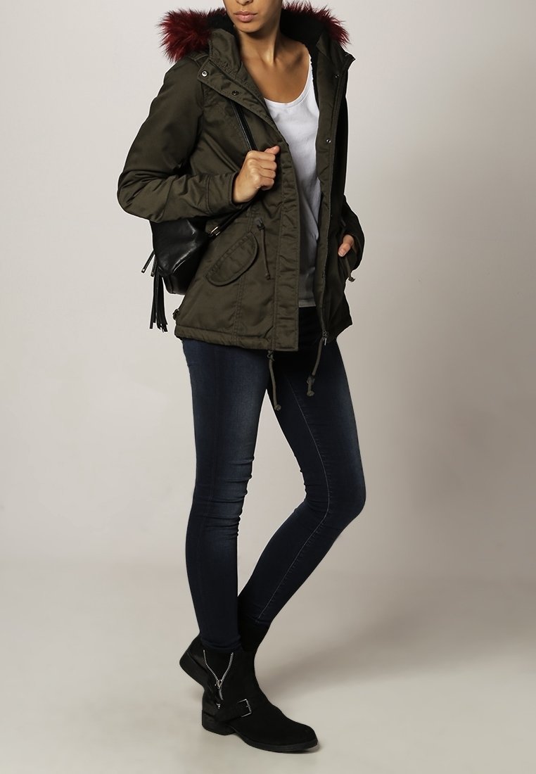 Kobieta ubrana w zieloną parkę z czerwonym futrzanym kapturkiem, białą koszulę, ciemne wąskie dżinsy, czarne botki za kostkę i nosząca czarny plecak.