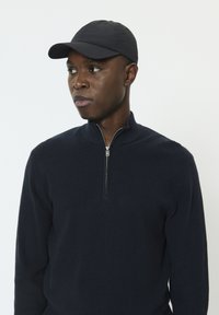 Pull bleu marine avec fermeture éclair et tissu texturé, associé à une casquette noire. Le pull a un col montant et des manches ajustées.