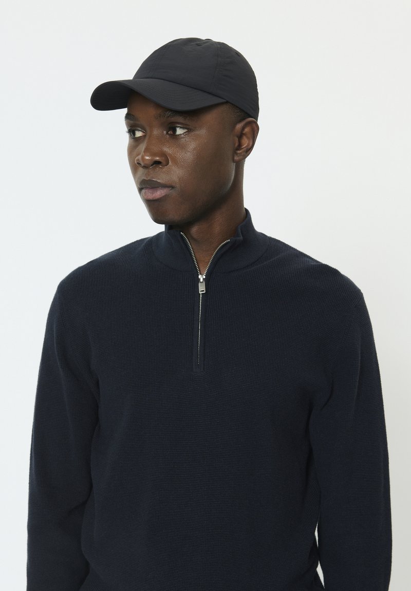 Pull bleu marine avec fermeture éclair et tissu texturé, associé à une casquette noire. Le pull a un col montant et des manches ajustées.