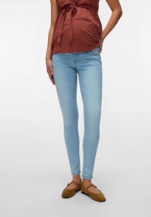 Femeie purtând blugi skinny bleu deschis, top maro legat și pantofi Mary Jane maro, stând cu o mână pe șold.