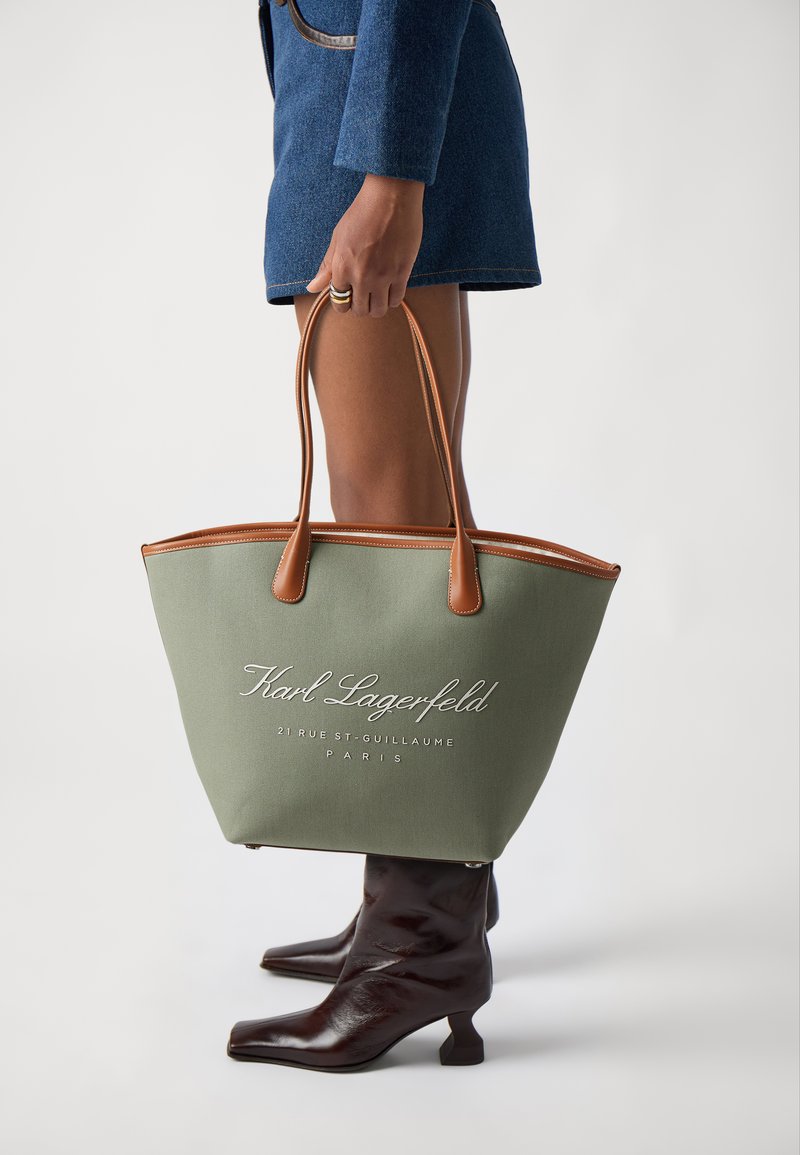 Grønn lerret tote bag med brune lærdetaljer, med hvit brodert tekst. Designet er åpent og romslig, med doble håndtak.