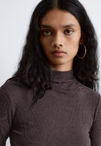 Langärmliger, dunkelvioletter Rollkragenpullover mit strukturiertem Paisleymuster. Das Model hat lange, wellige Haare und trägt große goldene Hoops.