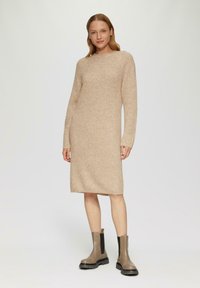 Beige gebreide jurk met een ronde hals, lange mouwen en geribbelde textuur; gecombineerd met bruine enkellaarsjes met zwarte elastische zijkanten.