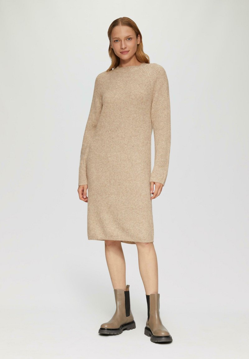 Beige gebreide jurk met een ronde hals, lange mouwen en geribbelde textuur; gecombineerd met bruine enkellaarsjes met zwarte elastische zijkanten.