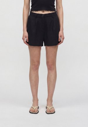 Personne portant un short noir sur mesure, un top court noir et des sandales beiges à bout carré, debout devant un fond clair uni.