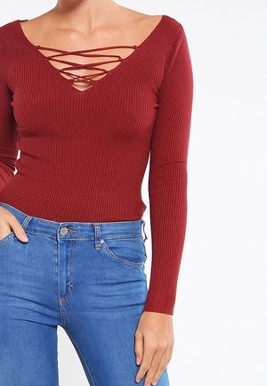 Woman wearing a fitted, long-sleeve, ribbed red top with crisscross laces at the neckline and high-waisted blue jeans.