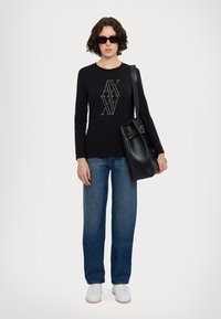 Camicia nera a maniche lunghe con logo grafico, jeans blu a gamba dritta, sneakers bianche e una borsa di pelle nera, abbinata a occhiali da sole scuri.