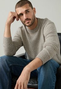 Lys grå strikket sweater med rund halsudskæring og rib-strikkede manchetter, parret med blå denim jeans, siddende på en sort læderstol.