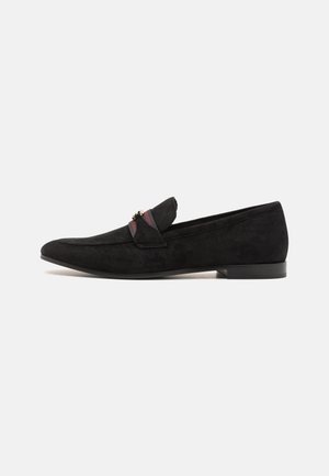 Zwarte suède loafer met lage hak, rood-zwart gestreepte stoffen riem en klein gouden metalen detail aan de zijkant.