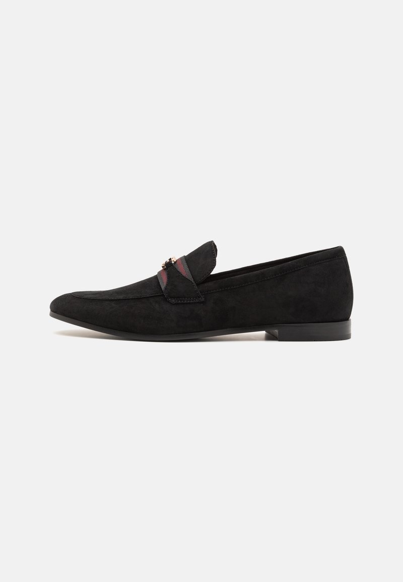 Mocassin en daim noir à talon bas, bride en tissu rayé rouge et noir, et petit détail en métal doré sur le côté.