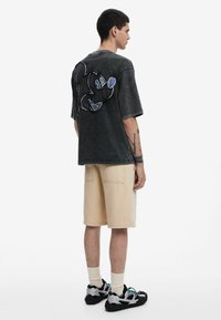 Camiseta de mezclilla negra de corte oversized con un gran diseño bordado de Mickey Mouse en la parte trasera. Combinada con pantalones cortos beige y zapatillas.