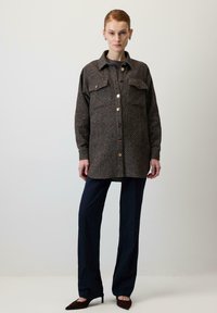 Kobieta ubrana w brązową, zygzakowatą, zapinaną na guziki kurtkę, granatowe, szerokie spodnie i ciemne szpilki w szpic, stojąca przy białej ścianie.