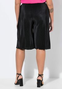 Culotte nere plissettate con orlo a scallop, caratterizzate da una texture lucida. Indossate con sandali neri con tacco, che mostrano gambe color pelle.