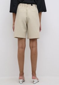 Shorts beige con un corte relajado, cinturilla elástica, dos bolsillos traseros y una textura suave, vistos desde atrás. Diseño sencillo.