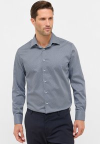 Man in een overhemd met lange mouwen en geometrisch patroon, in donker gekleurde broek, die iets naar de zijkant kijkt tegen een effen achtergrond.