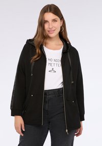 Giacca nera con zip e cappuccio, caratterizzata da un tessuto testurizzato sulle maniche e sulle tasche. Indossata sopra una maglietta bianca con grafica e pantaloni scuri.