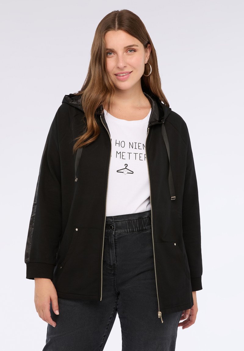Giacca nera con zip e cappuccio, caratterizzata da un tessuto testurizzato sulle maniche e sulle tasche. Indossata sopra una maglietta bianca con grafica e pantaloni scuri.