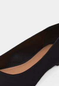 Γυναικείο μαύρο χαμηλό παπούτσι με στρογγυλή μύτη και μπεζ εσωτερική σόλα σε λευκό φόντο.