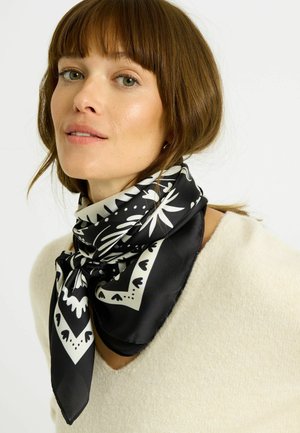 Mujer con cabello castaño claro lleva una bufanda blanca y negra con estampado atada alrededor del cuello y un suéter color crema, mirando ligeramente hacia la izquierda.