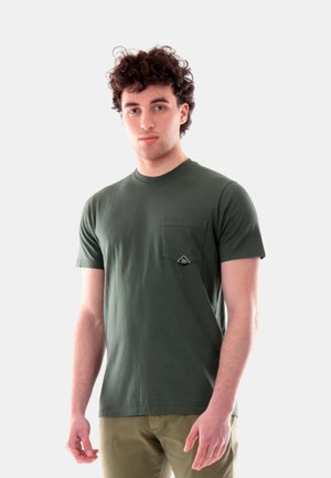 Giovane uomo con capelli ricci che indossa una maglietta verde oliva con taschino sul petto e pantaloni beige su uno sfondo semplice.