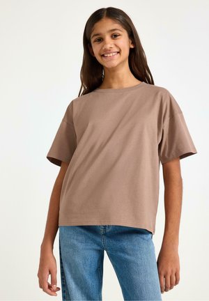 Smilende pige iført en løs taupe kortærmet skjorte og blå jeans mod en ensfarvet hvid baggrund.