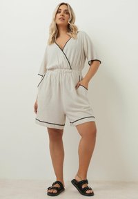 Beige linnéromper med korta ärmar, omslagdesign, svarta kantdetaljer, elastisk midja och fickor. Bärs med svarta utsmyckade sandaler.