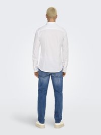 Personne aux cheveux blonds courts portant une chemise blanche ajustée à manches longues, un jean bleu et des chaussures beiges, debout de dos sur un fond uni.