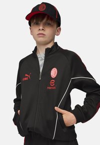 Chaqueta deportiva negra con cremallera y detalles en blanco, logo rojo y bordado, combinada con una gorra negra con logo rojo. Estilo con una camiseta gris por debajo.