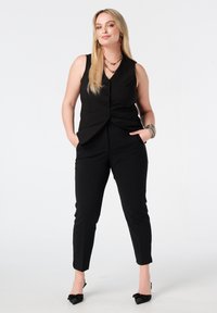 Zwarte vest met knopen gecombineerd met zwart, op maat gemaakte broek, met een slanke pasvorm en enkellengte. Model gestyled met sieraden en hakken.