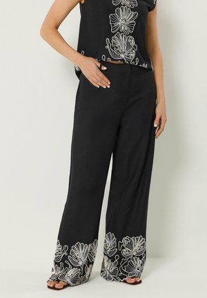Mujer que lleva pantalones negros de pierna ancha con bordado floral blanco en los bajos y una camiseta negra a juego con un diseño floral similar.
