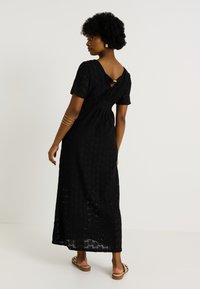 Femme avec des cheveux afro, portant une robe maxi en crochet noire à manches courtes et des sandales à imprimé léopard, se tenant contre un fond blanc.
