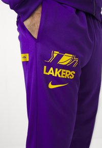 Calças de treino roxas com o logótipo dos "Lakers" em amarelo, juntamente com o símbolo da Nike em amarelo. Textura suave com bolsos laterais.