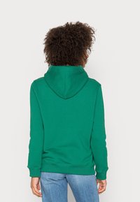 Sudadera verde con una textura suave, que presenta una capucha con costura cruzada, puños acanalados y dobladillo, vista desde la parte de atrás sobre un fondo liso.