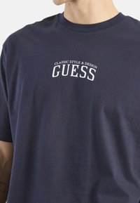 Camiseta de algodón azul marino con mangas cortas y cuello redondo, con "ESTILO CLÁSICO Y DISEÑO GUESS" impreso en blanco.