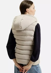 Beżowa kamizelka puchowa z kapturem, z pikowanymi sekcjami i miękką teksturą. Nosi się na ciemnej bluzce z długim rękawem, z widocznymi szwami na plecach.